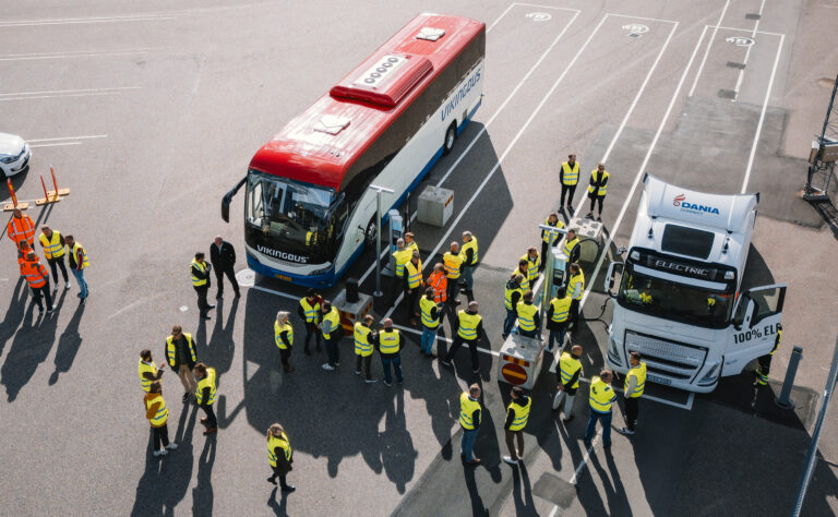 Laddstation med två laddande fordon, buss och lastbil. Folksamling klädda med varselvästar mellan fordonen. Staket intill containerterminal med uppställda containrar i bakgrunden.